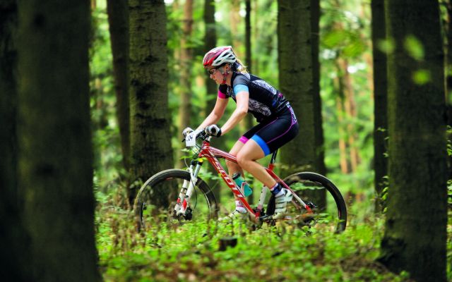 Kobieta w profesjonalnym stroju kolarskim pokonuje na rowerze leśną trasę terenową. Do kierownicy ma przypięty numer startowy. Wokół drzewa.